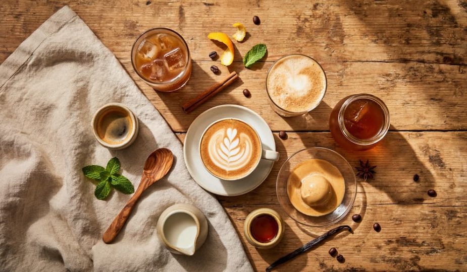 Vue du dessus d'une table en bois avec six boissons café différentes : expresso, café glacé, latte art, frappé, affogato et cold brew, entourés de grains de café, épices et ustensiles, ambiance boho estivale