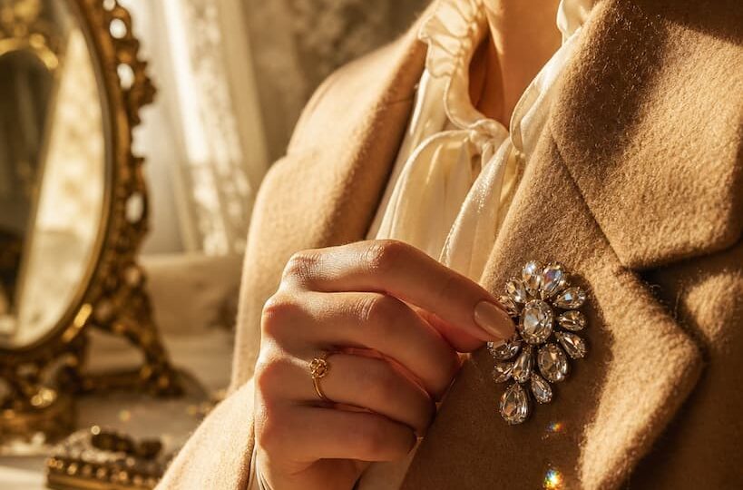 Main élégante fixant une broche vintage à strass scintillants sur le revers d'un manteau en laine camel, ambiance boudoir doré